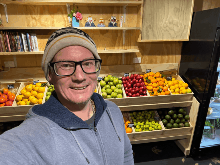 A white man wearing glasses and a beanie stands smiling in front of several bins of colorful fruit.